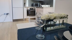 Eine moderne Küche mit weißen Schränken, einer Mikrowelle, einem Backofen und einem kleinen Kühlschrank. Ein Esstisch mit blauer Tischdecke, zwei Weingläsern und einer dekorativen Schale mit weißen Blumen.