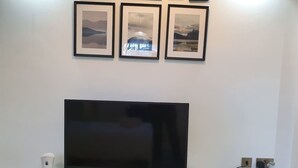 Ein Flachbildfernseher auf einem weißen Ständer, mit Fernbedienung, einem weißen Lautsprecher und drei gerahmten Bildern über der Wand.