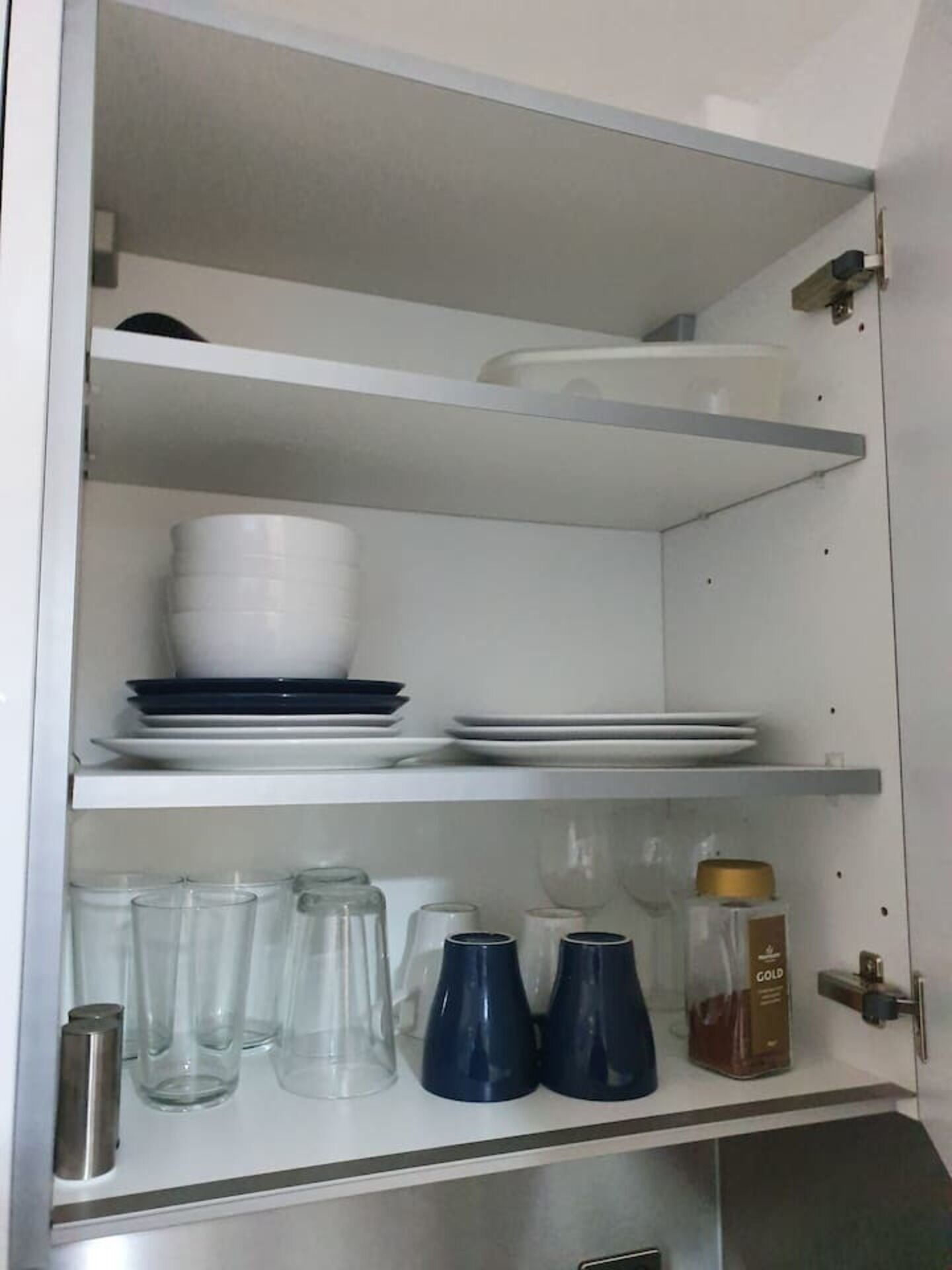 A kitchen cabinet with shelves holding plates, bowls, glasses, and a bottle of gold syrup.