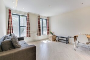 Un salon moderne avec un canapé, une petite table et un fauteuil. Il y a deux fenêtres avec des rideaux, un radiateur et une plante posée sur une table d’appoint.