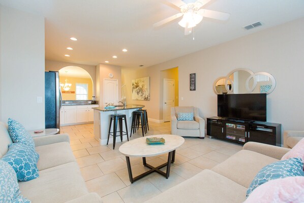 A modern living room with a sofa, a coffee table, and a TV stand. There is a kitchen area in the background with a bar stool and a refrigerator.