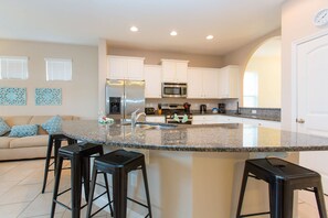 A modern kitchen with white cabinets, a stainless steel refrigerator, a microwave, and a granite countertop. There are two black bar stools and a white door in the background.