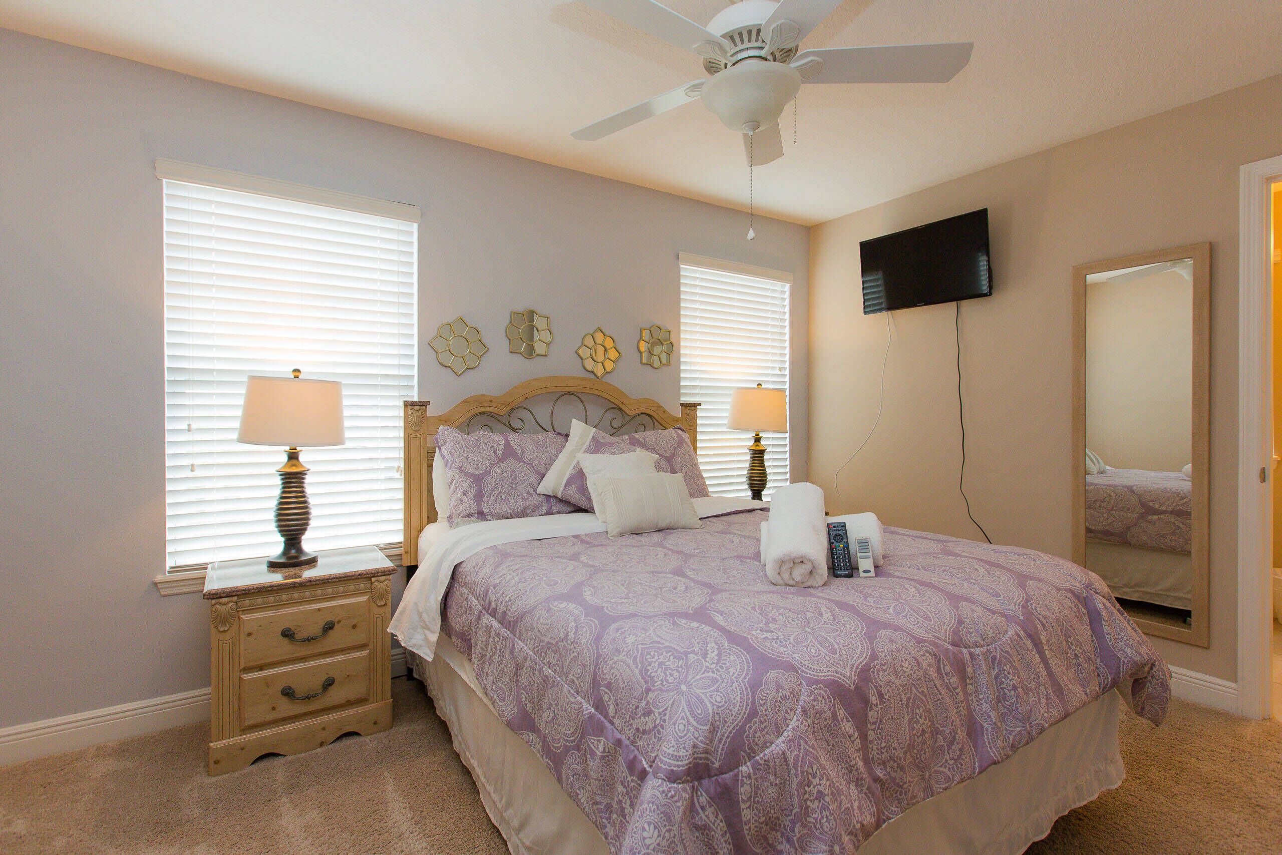 A bedroom with a bed, two bedside tables with lamps, a ceiling fan, a TV mounted on the wall, and a large mirror.
