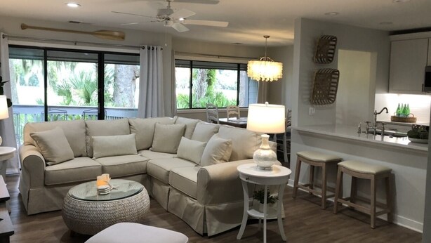 A modern living room with a sectional sofa, a round coffee table, a ceiling fan, and a kitchen area with a bar stool.