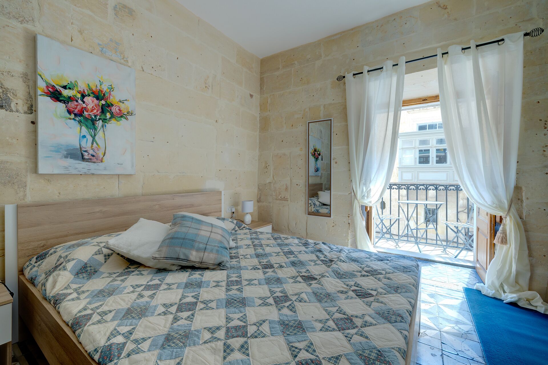 A bedroom with a bed, a painting, a mirror, and a view of a balcony.