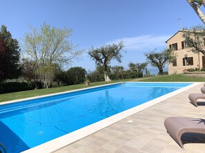 A swimming pool with a tiled deck, surrounded by greenery and a building with a Mediterranean architectural style.
