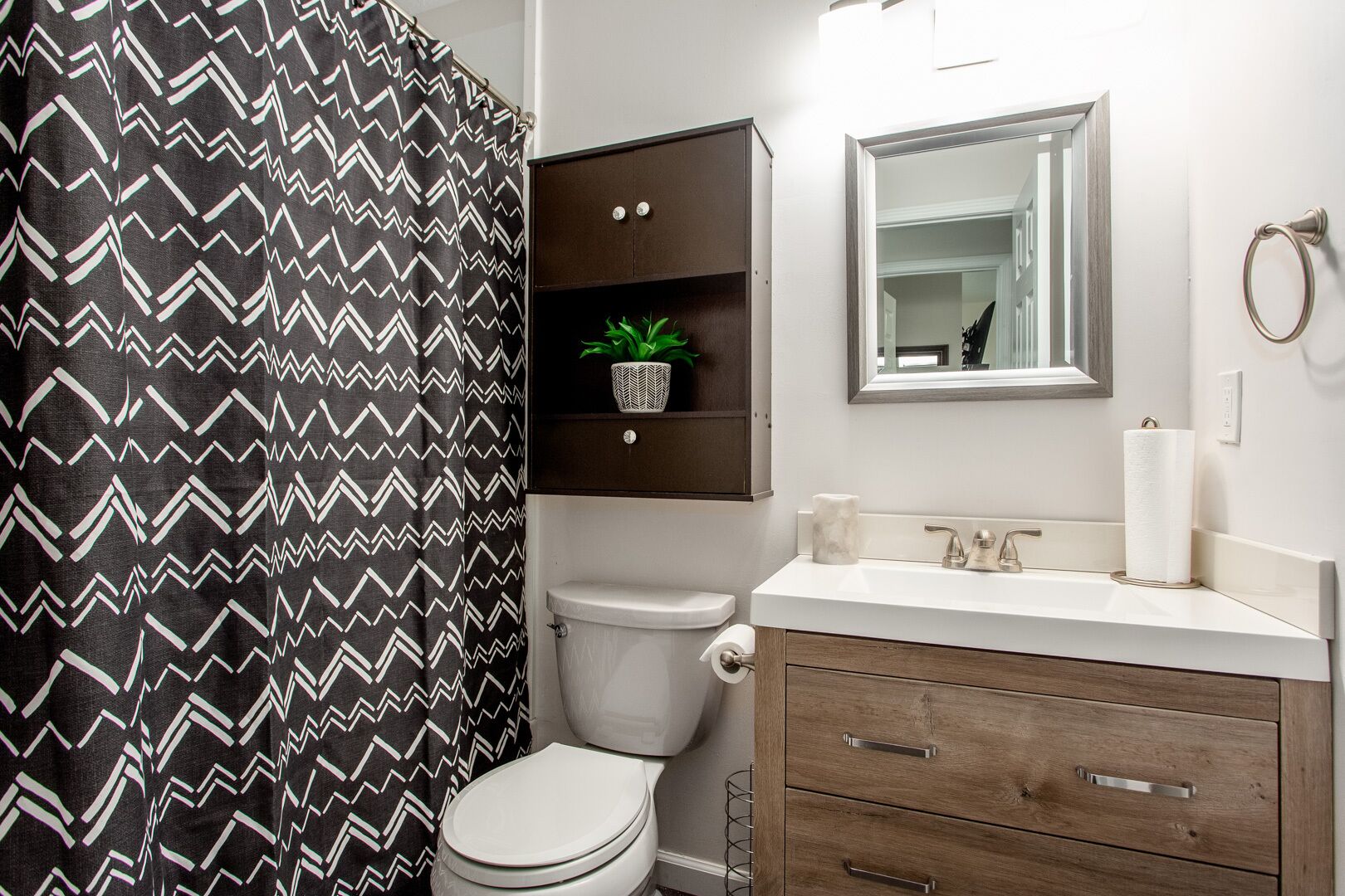 A bathroom with a black and white zigzag shower curtain, a white toilet, a wooden vanity with a mirror, and a small potted plant on the shelf.