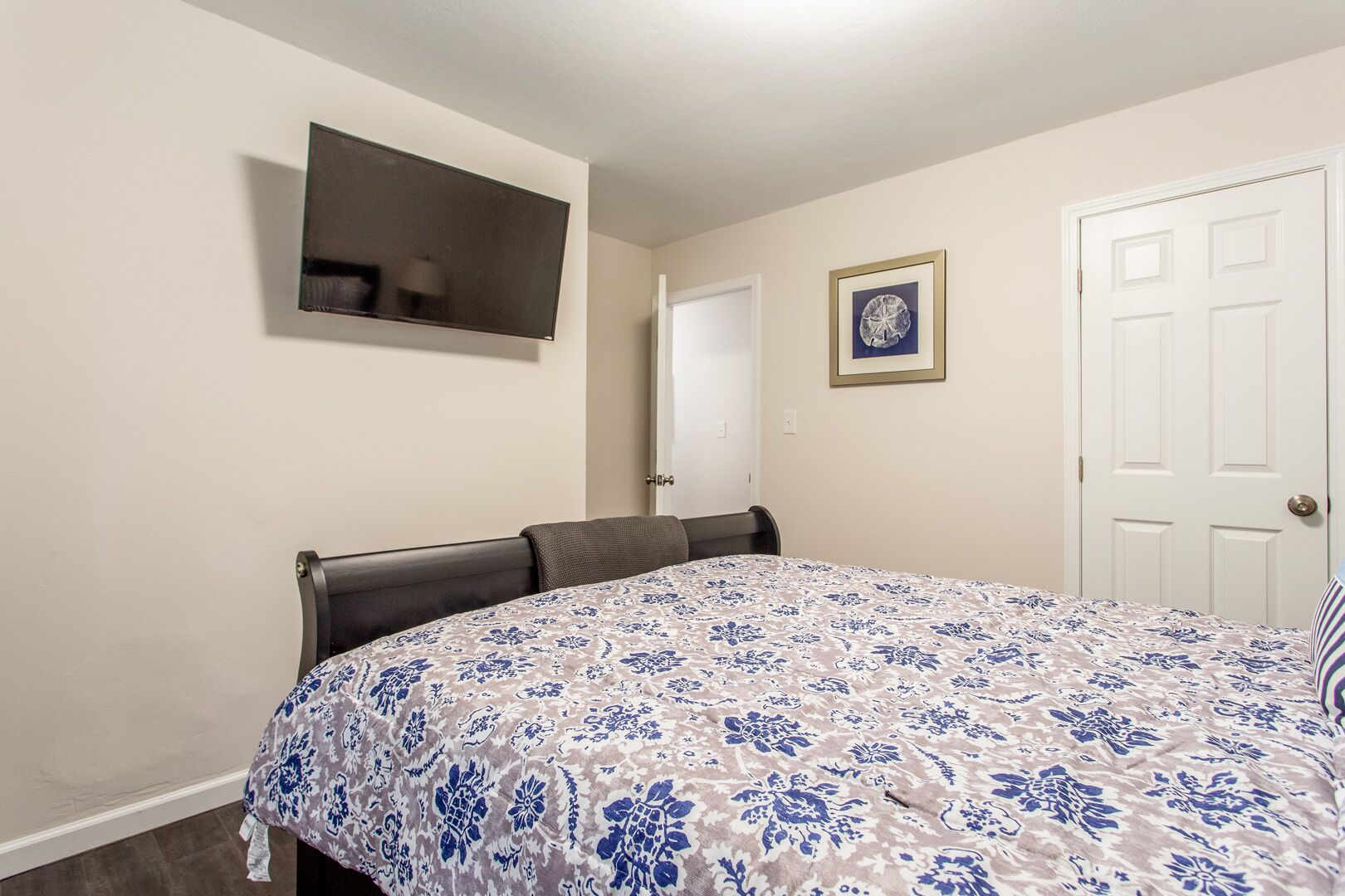 Un dormitorio con una cama, un televisor montado en la pared, una puerta y un cuadro enmarcado.