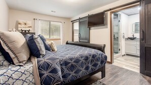 A bedroom with a bed, a TV, a sliding door to a bathroom, and a window with blinds. - Stunning Custom Ranch in Central Roswell (Roswell)