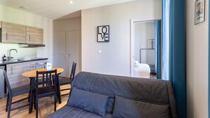 Ein modernes Wohnzimmer mit grauem Sofa, einem Holzesstisch mit Stühlen, einer Küchenzeile mit Schränken und Spüle sowie einem durchgegangenen Türrahmen einsehbaren Schlafzimmer.