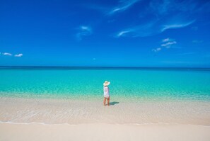 A person standing on a sandy beach with clear turquoise water and a clear blue sky.