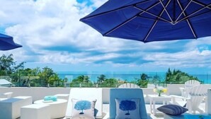 A rooftop lounge with white furniture, blue cushions, and a view of the ocean.