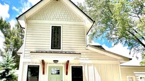 Een woonhuis met twee verdiepingen, een puntig dak, een veranda met een groene deur en een raam met decoratieve roedeversiering.