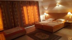 A hotel room with a double bed, a single bed, a nightstand with a lamp, a small table, and a patterned rug. - Hotel Sindbad Center Sousse (Sousse)
