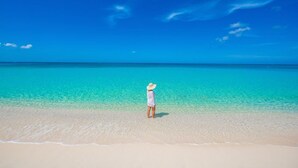 A person standing on a sandy beach with clear turquoise water and a clear blue sky. - 7 Mile Beach private Beach Club, ocean views from Roof deck and Pool Pavilion (West Bay)