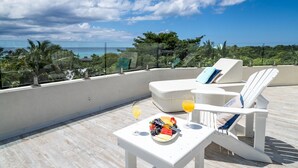 A rooftop terrace with two white lounge chairs, a small table with a fruit platter and drinks, and a view of the ocean and palm trees. - 7 Mile Beach private Beach Club, ocean views from Roof deck and Pool Pavilion (West Bay)