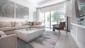 A modern living room with a sectional sofa, a glass coffee table, and a view of the outdoors through large sliding glass doors. - 7 Mile Beach private Beach Club, ocean views from Roof deck and Pool Pavilion (West Bay)