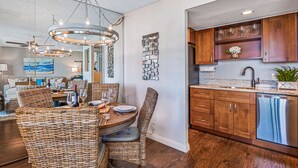 Un coin repas avec des chaises en osier et une table ronde, une cuisine avec des meubles en bois et un lave-vaisselle, et un ventilateur de plafond.