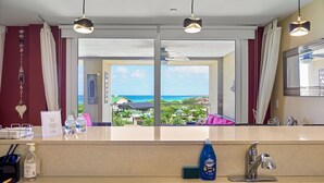 A hotel room with a large window offering a view of the sea and buildings, a sink with a mirror, and a counter with a welcome sign and amenities. - OUTSTANDING OASIS 4th FLOOR CONDO At EAGLE BEACH (Oranjestad, Aruba)