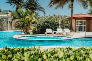 A swimming pool with two white lounge chairs, surrounded by palm trees and a small building with a tiled roof.