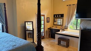 A bedroom with a four-poster bed, a small stool, a desk with a chair, a sink, and a window with curtains.