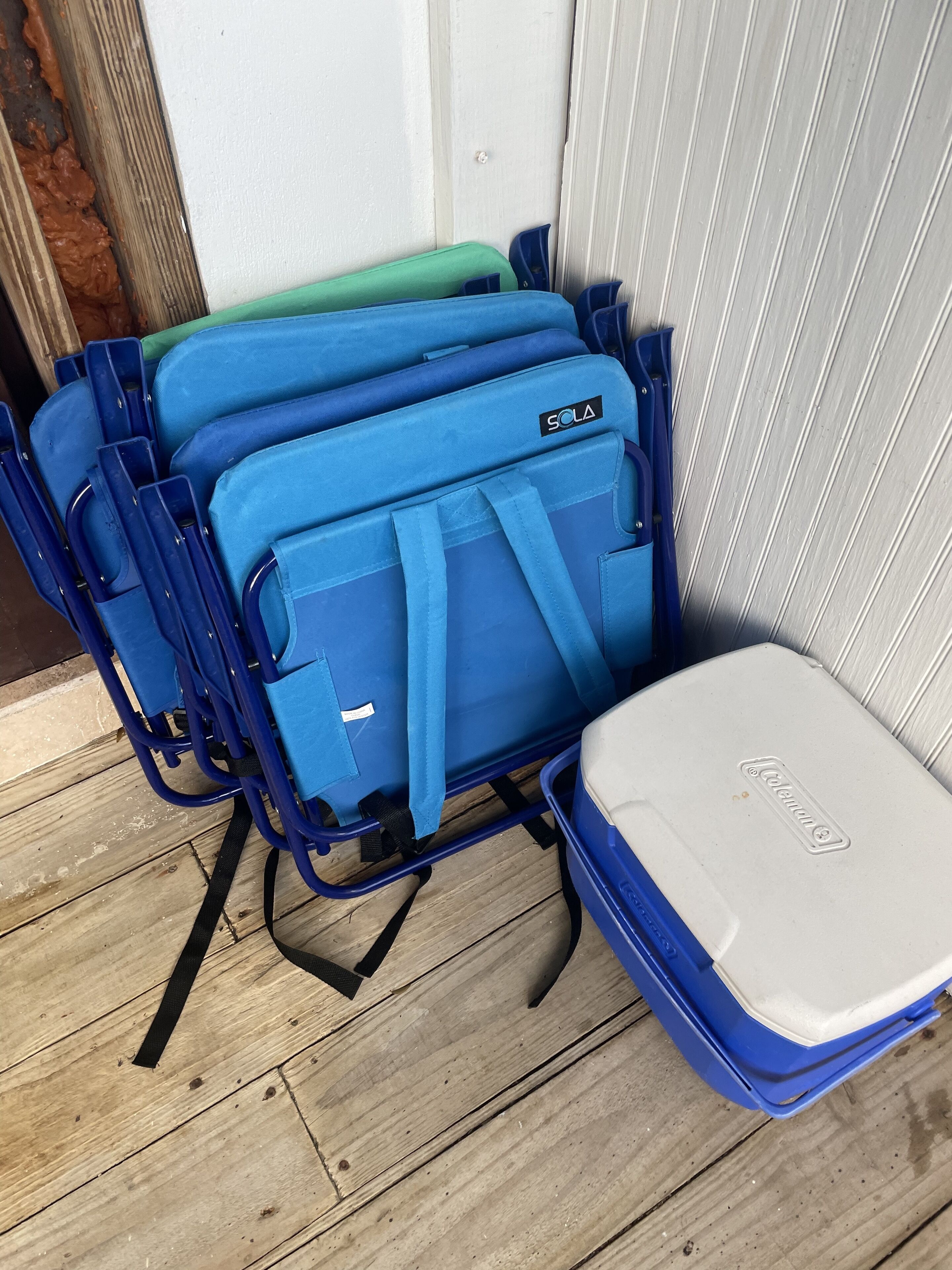 A stack of blue and green suitcases with a Coleman brand cooler on a wooden floor.