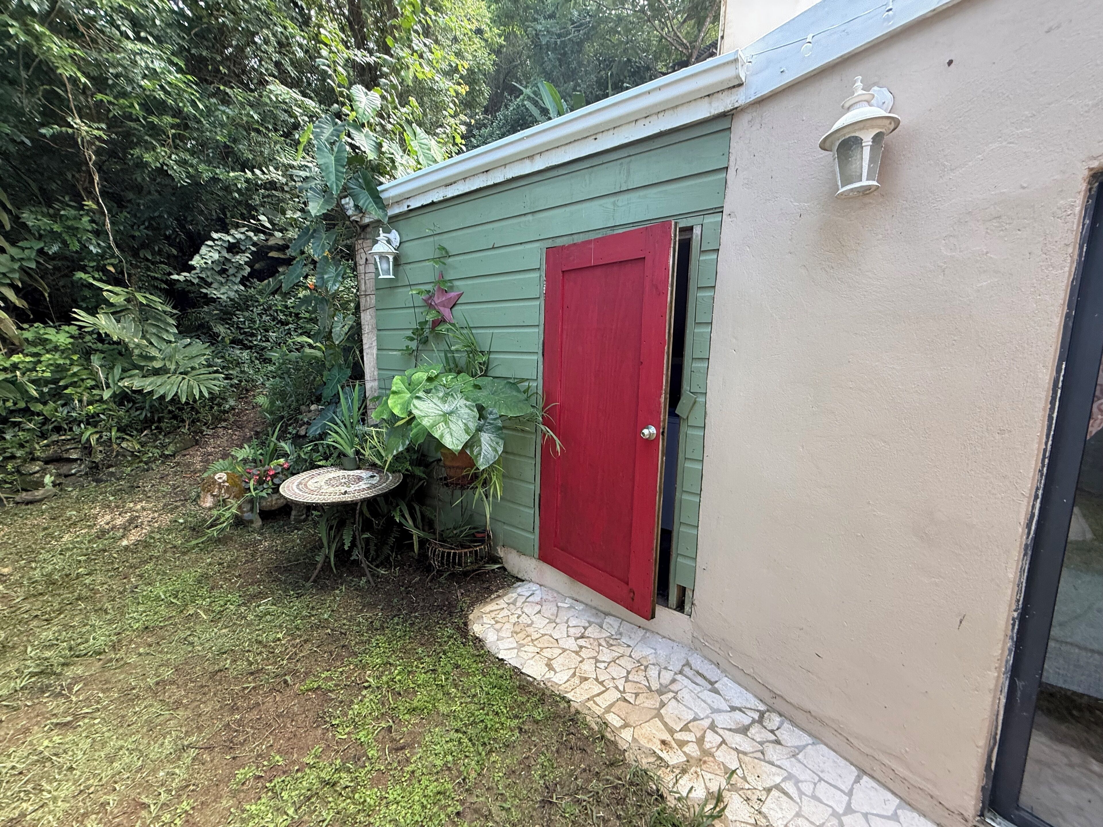 Un petit abri de jardin vert avec une porte rouge, entouré d’une végétation luxuriante et agrémenté d’une petite table ronde sur laquelle sont posées des plantes.