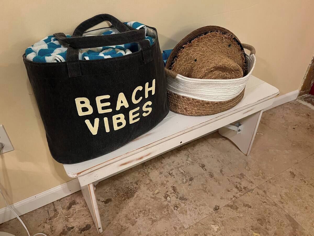Una bolsa de tela negra con el texto "BEACH VIBES" y una cesta tejida sobre un estante blanco.