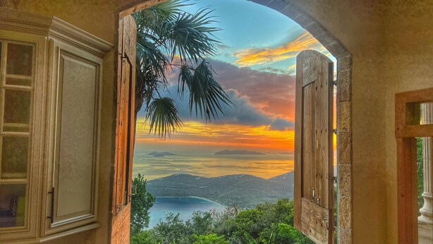 A room with an open wooden door leading to a balcony with a view of a sunset over a body of water and hills.
