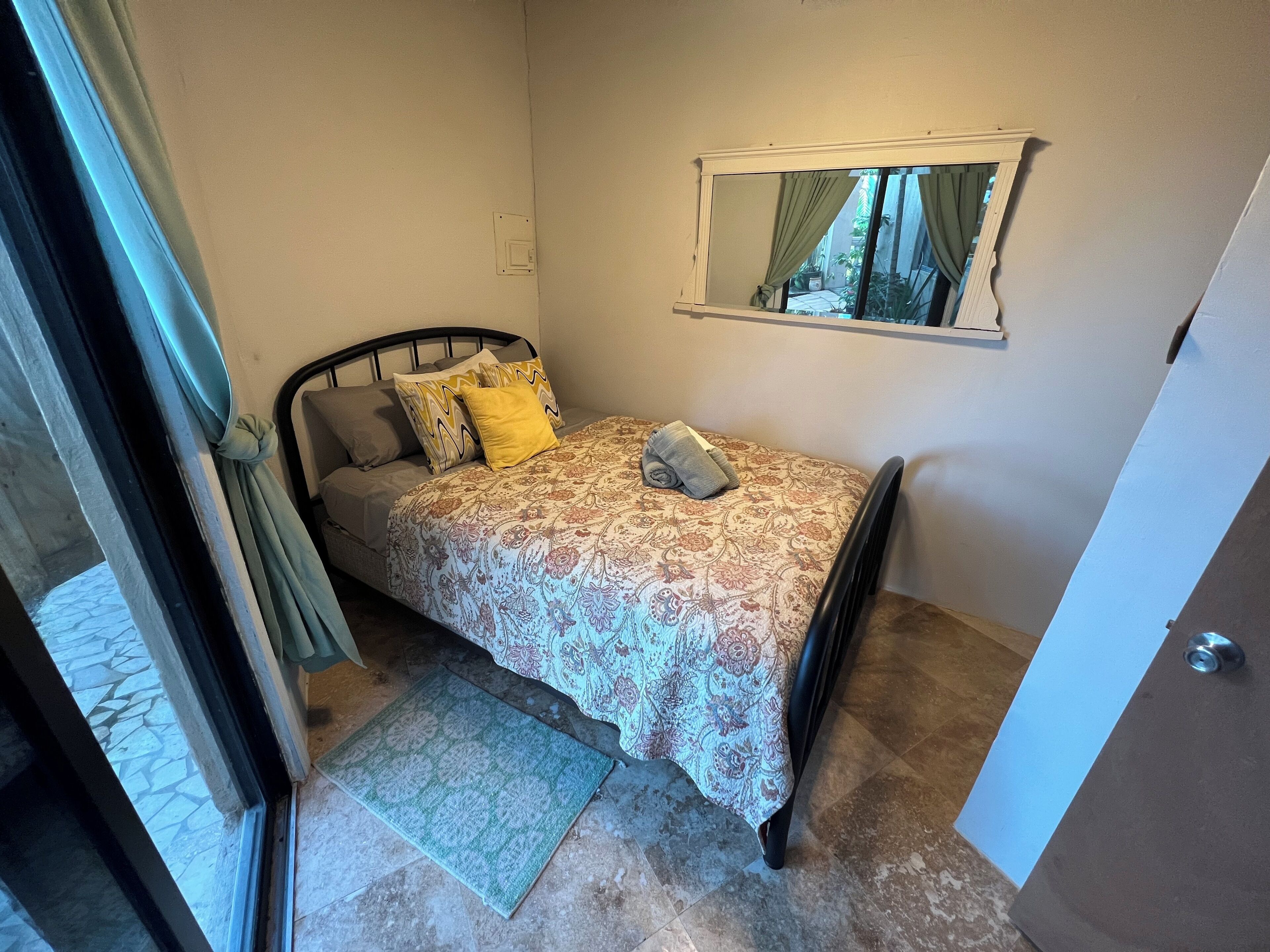 Un dormitorio pequeño con cama, mesita de noche, espejo y ventana con cortinas.