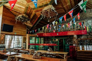 Interior r ústico de madera con bancos y mesas de madera, una pared de piedra y un mostrador rojo.