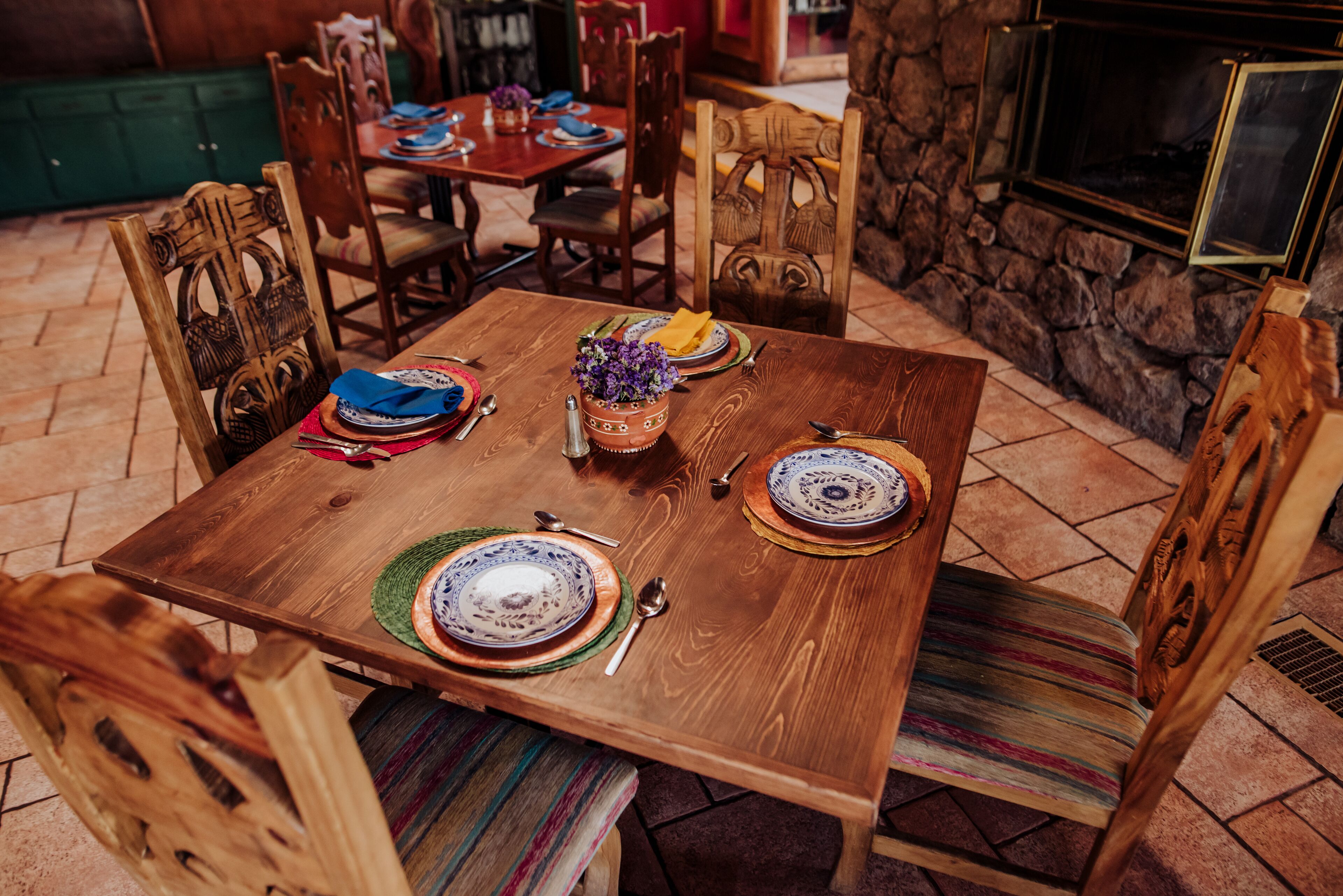Una mesa de madera para comer dispuesta con platos, cubiertos y un jarrón de flores moradas.
