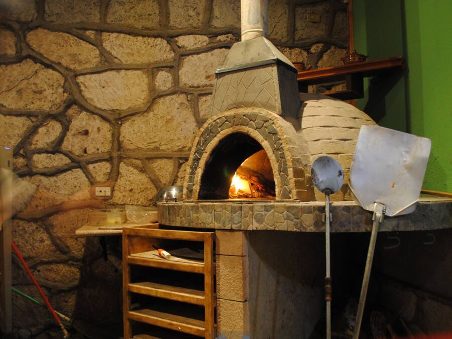 A stone-built brick oven with a chimney, a wooden ladder, and a metal pizza peel.