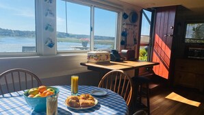 A dining area with a table set for breakfast, a view of the water, and a kitchen area visible through an open door. - Wellfleet Harbor Waterfront-Duck Creek Cottage (Wellfleet)