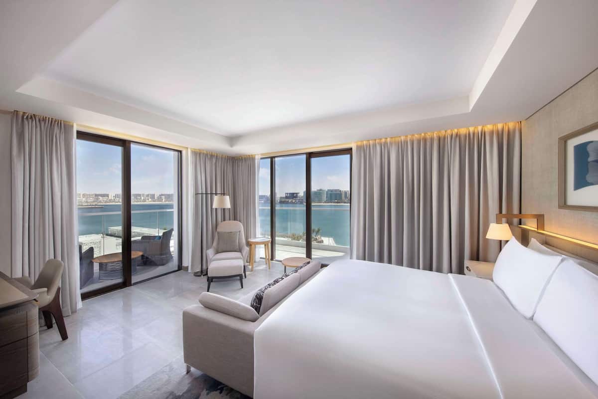 Ein modernes Hotelzimmer mit einem großen Bett, einer Couch, einem Sessel, einem kleinen Tisch und Blick auf die Stadt und das Wasser durch große Fenster.