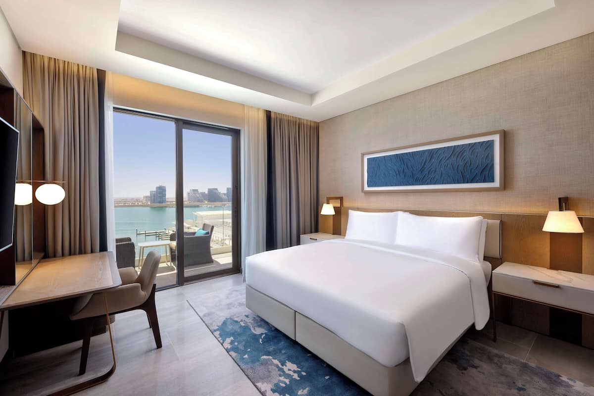 Ein Hotelzimmer mit einem großen Bett, einem Schreibtisch und einem Stuhl. Durch das Fenster bietet sich ein Blick auf die Stadt und das Wasser.