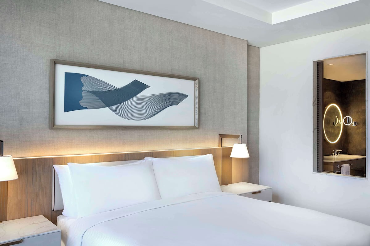 Ein modernes Hotelzimmer mit einem großen Bett, zwei Nachttischen mit Lampen, einem an der Wand hängenden abstrakten Kunstwerk und einem Spiegel mit Neonbeleuchtung.