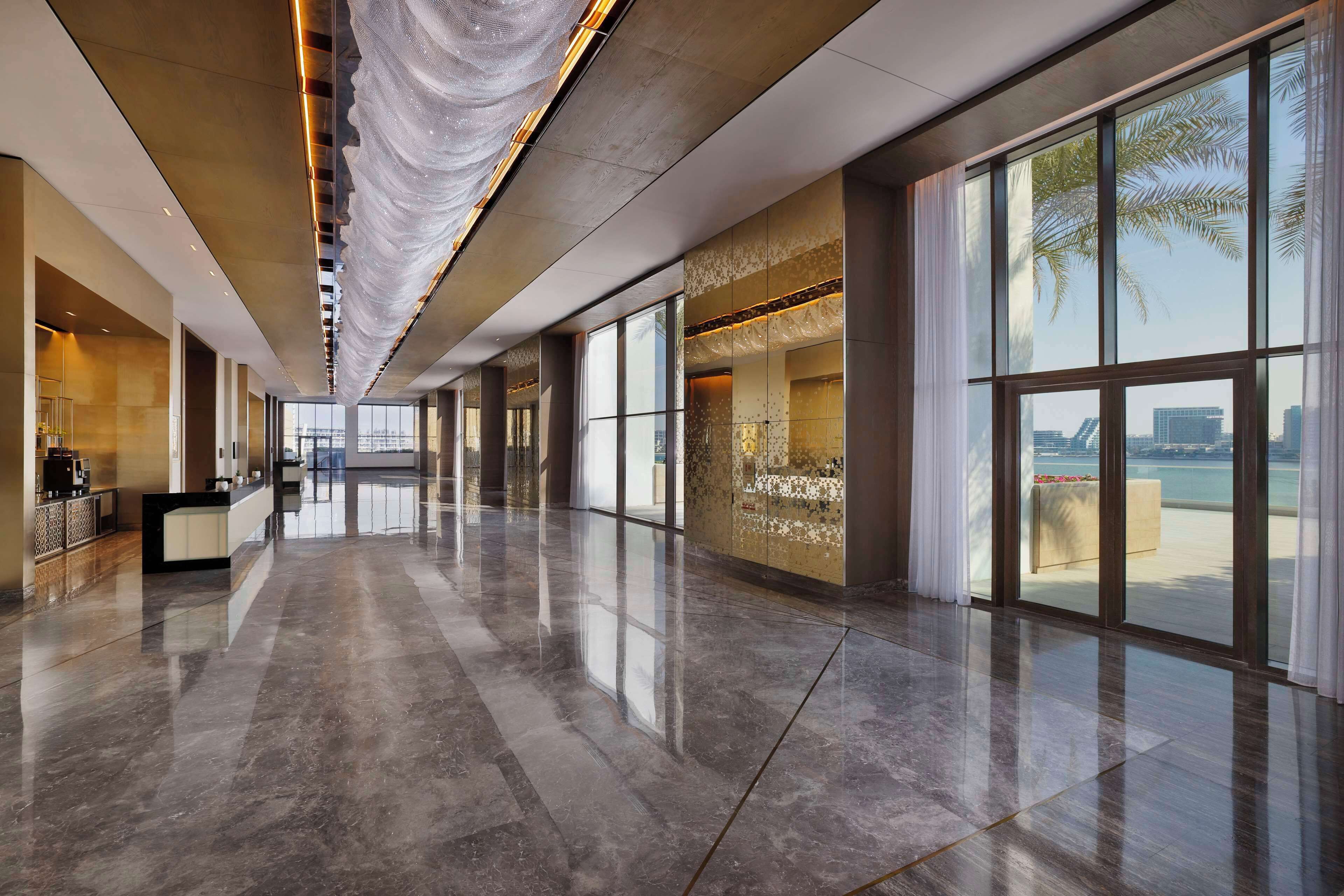 A spacious, modern hotel lobby with a reflective marble floor, large windows offering a view of the sea, and a reception area with a counter and seating.