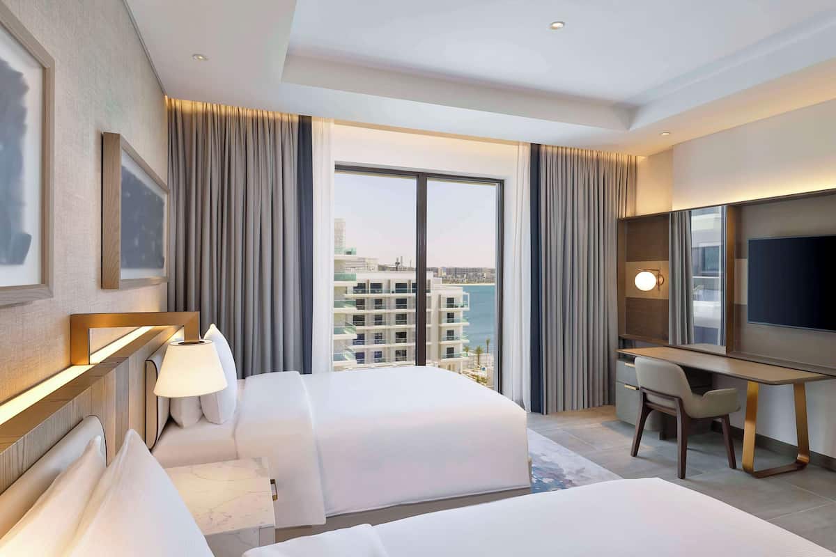 Ein Hotelzimmer mit zwei Betten, einem Schreibtisch, einem Sessel, einem Fernseher und einem großen Fenster mit Blick auf die Stadt.