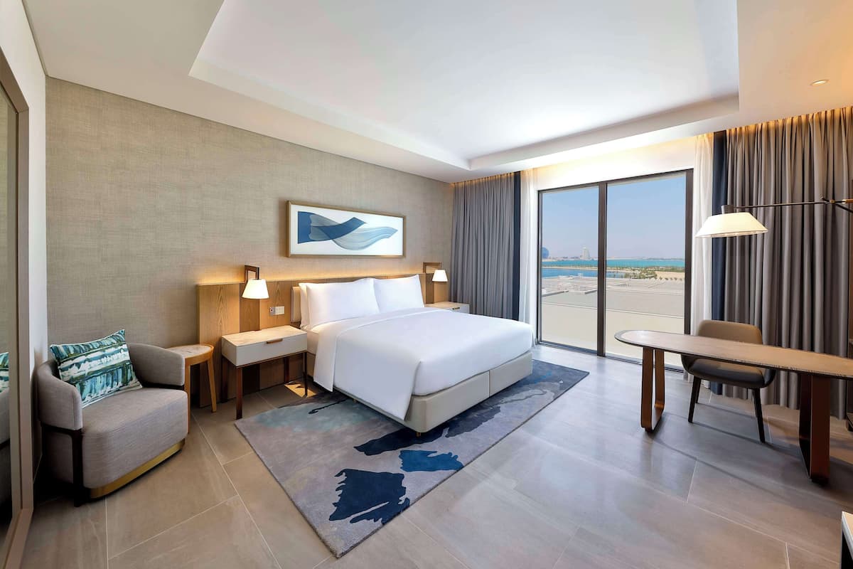Ein modernes Hotelzimmer mit einem großen Bett, einem Schreibtisch, einer Sitzecke und Blick auf den Strand durch das Fenster.