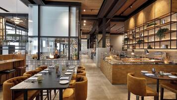 A modern restaurant with a long dining area, a kitchen visible through glass walls, and a bar area with a wooden counter.