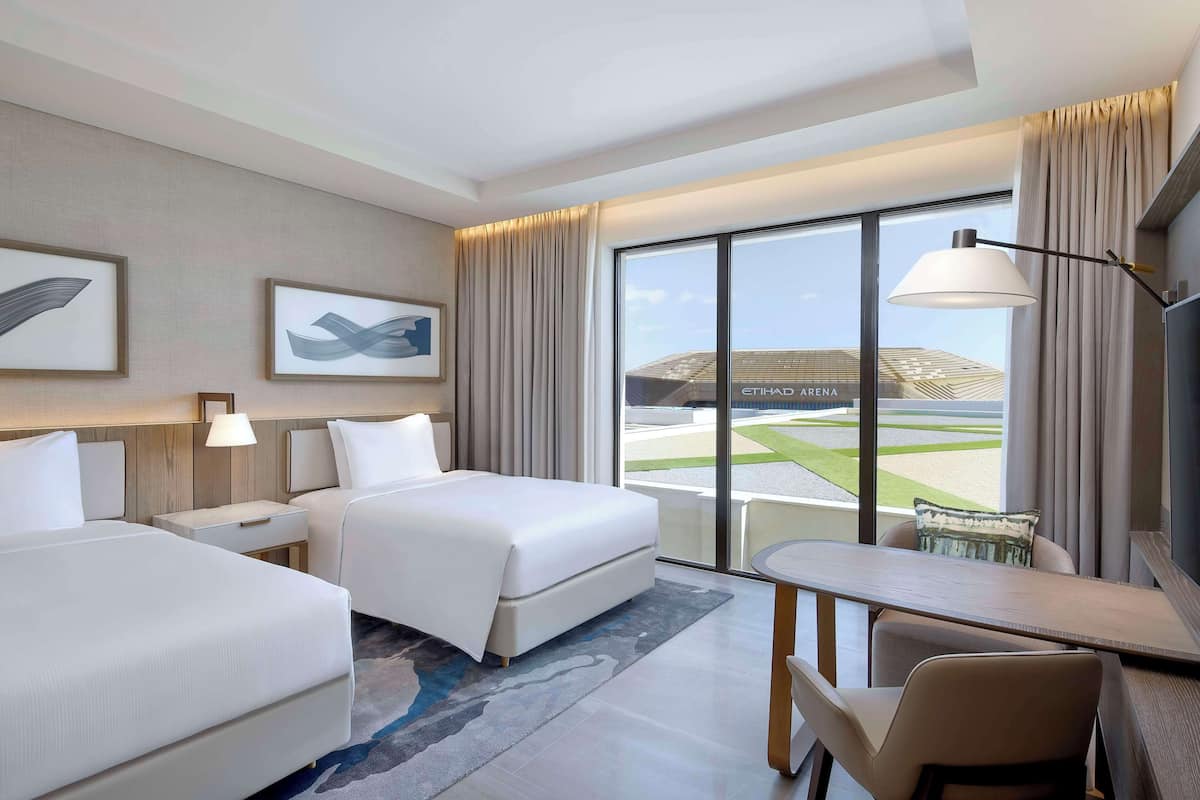 Ein Hotelzimmer mit zwei Betten, einem Schreibtisch, einem Stuhl, einer Lampe und einem großen Fenster mit Blick auf ein Gebäude.