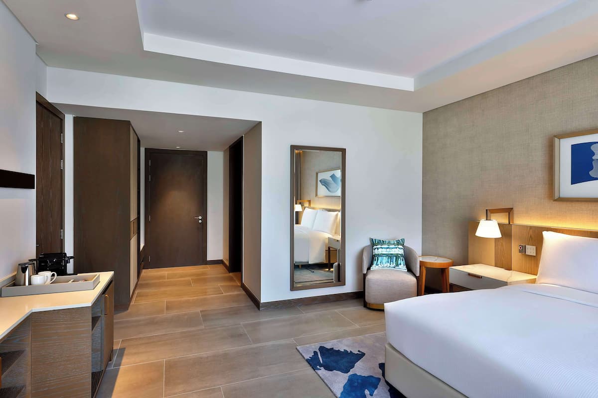 Ein modernes Hotelzimmer mit Bett, Nachttisch, Lampe und Spiegel. Es gibt eine kleine Sitzecke mit Stuhl und Tisch. Der Raum hat einen Holzboden und eine helle Wand.
