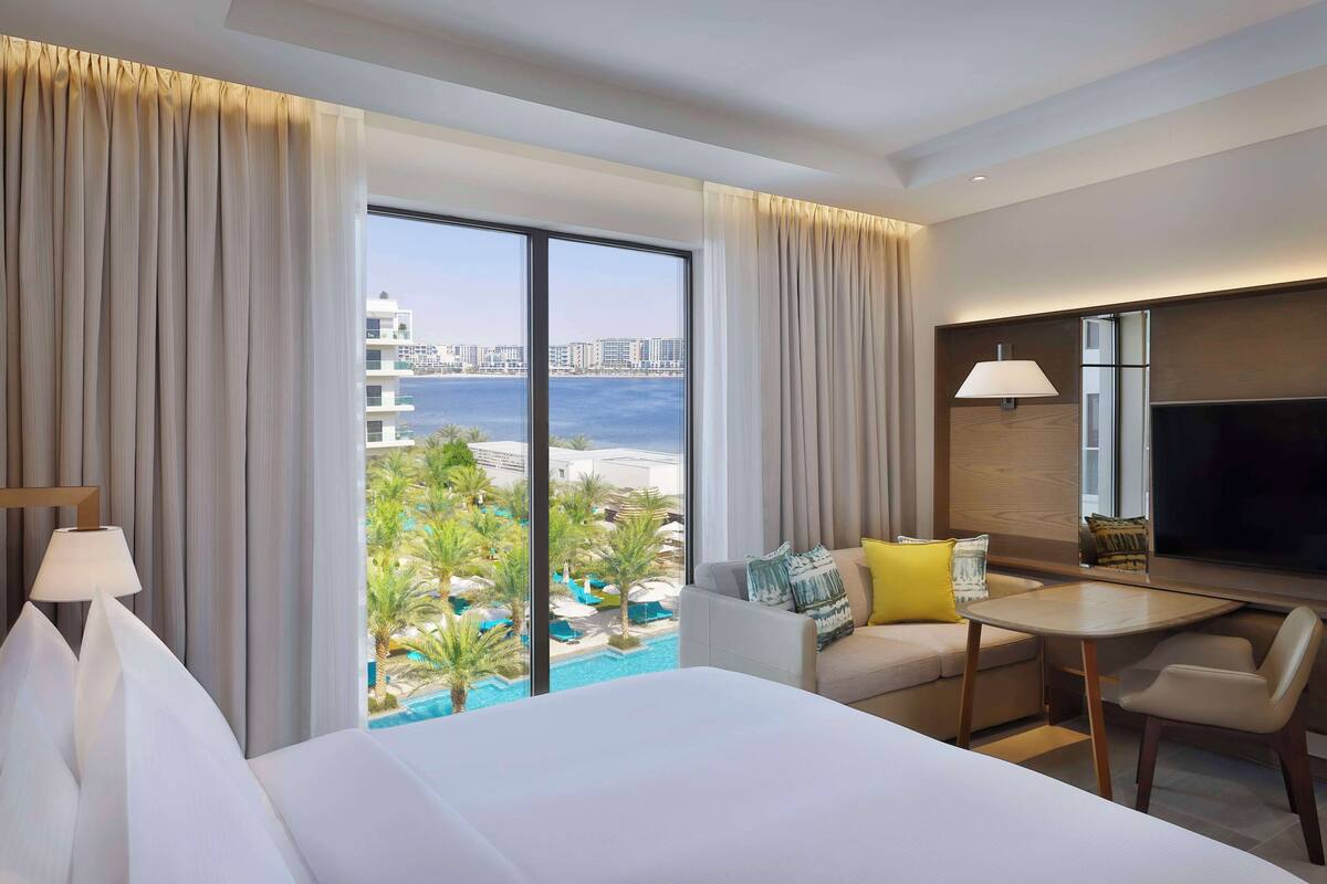 Ein Hotelzimmer mit einem großen Bett, einem Schreibtisch, einem Sessel, einem Fernseher und Blick auf einen Pool und Gebäude.