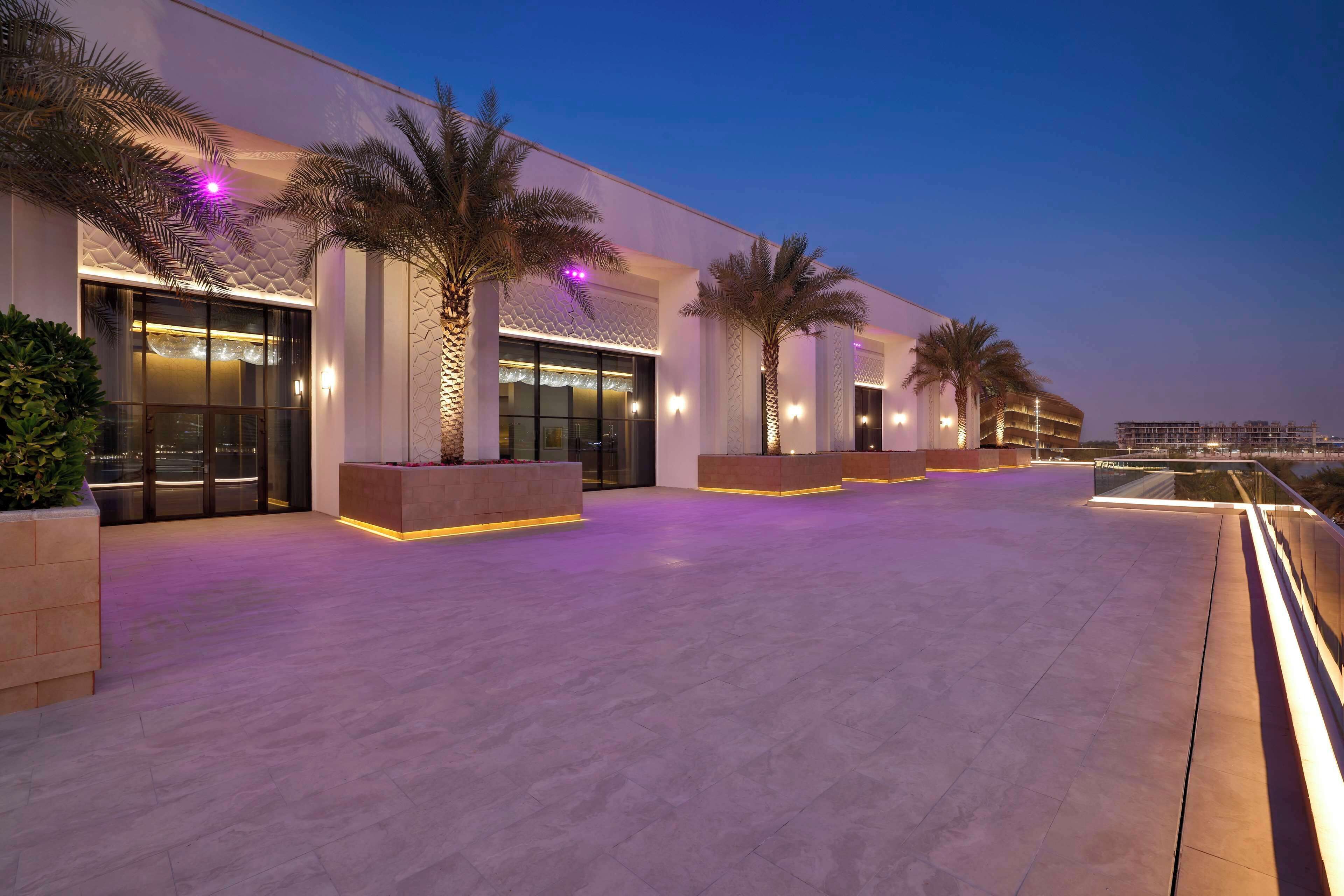 Un hôtel moderne avec une façade élégante, un vaste espace extérieur, des palmiers et des entrées illuminées.
