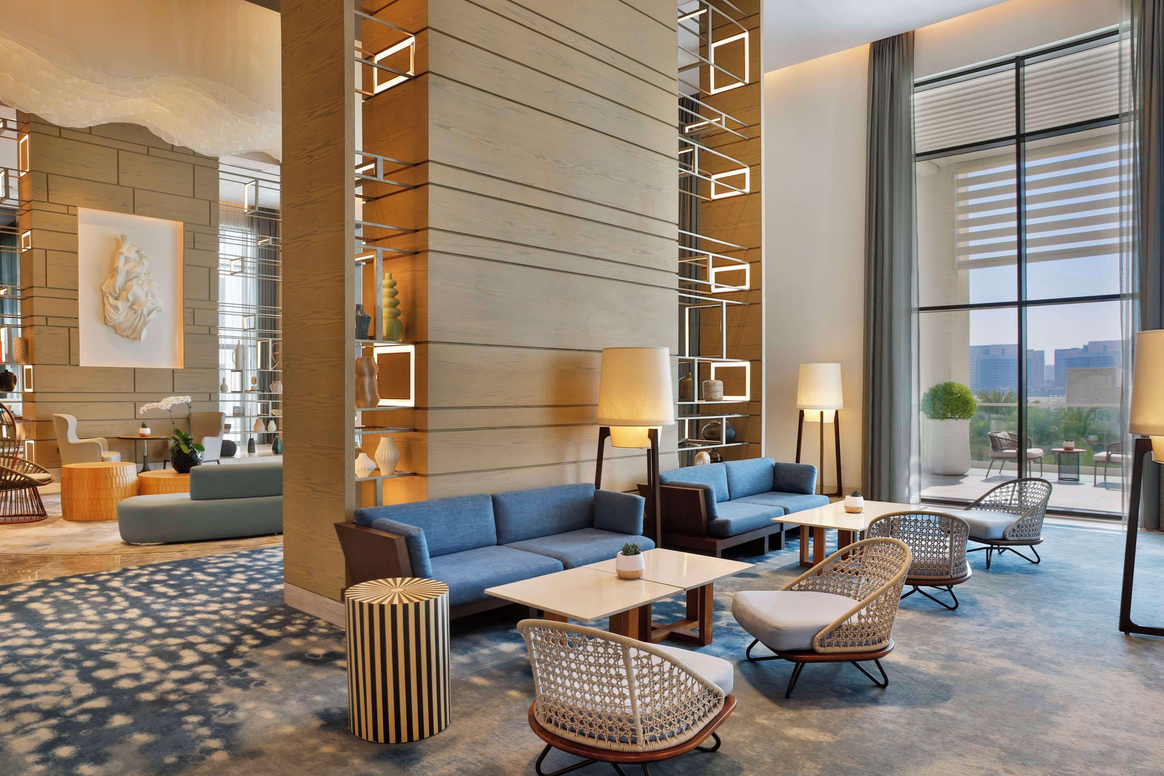 Een moderne hotellobby met een blauwe bank, rieten stoelen, een bijzettafeltje en een groot raam met uitzicht op de stad.