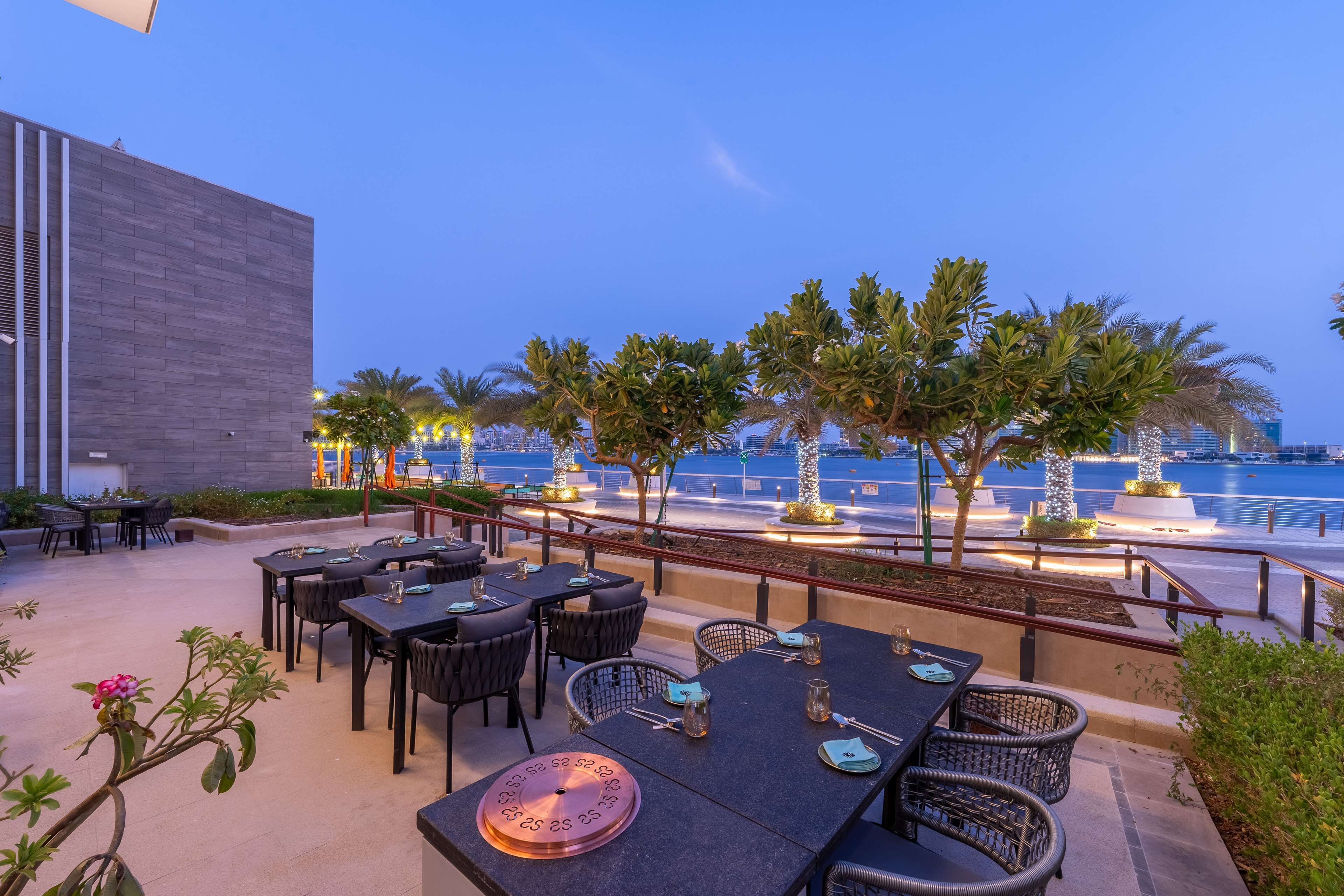 Un área de comedor al aire libre con mesas y sillas, palmeras y vista al agua.