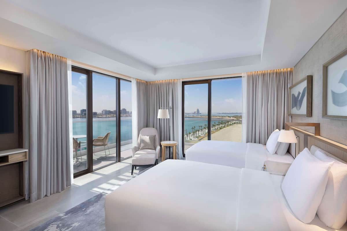 Ein Hotelzimmer mit zwei Betten, einem Sessel, einem kleinen Tisch und einem großen Fenster mit Blick auf das Meer und die Stadt.