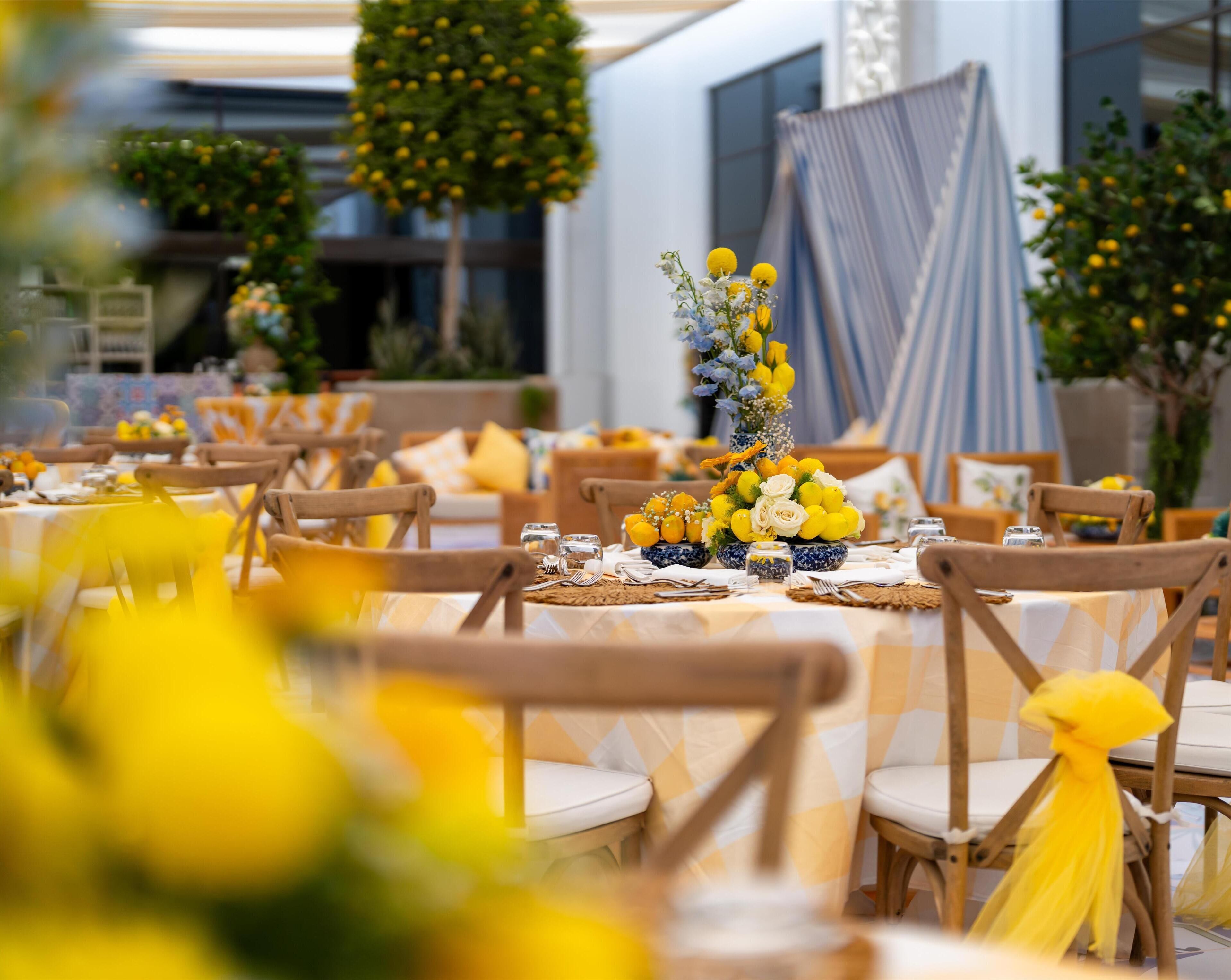A dining area with round tables set for a meal, featuring yellow and white tablecloths, wooden chairs, and a central floral arrangement.