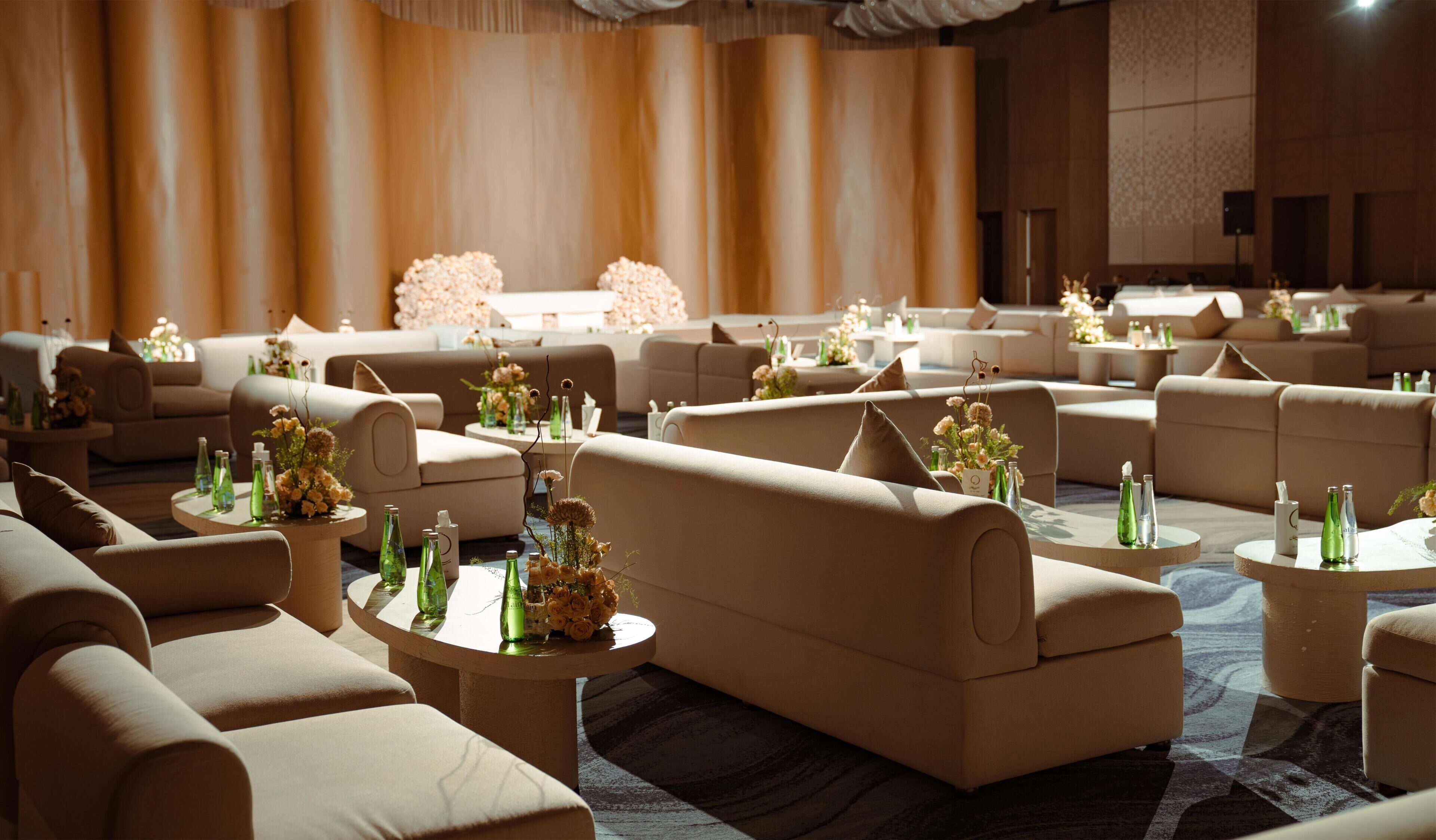 A spacious hotel lobby with multiple seating areas, including sofas and round tables, adorned with floral arrangements and green bottles.